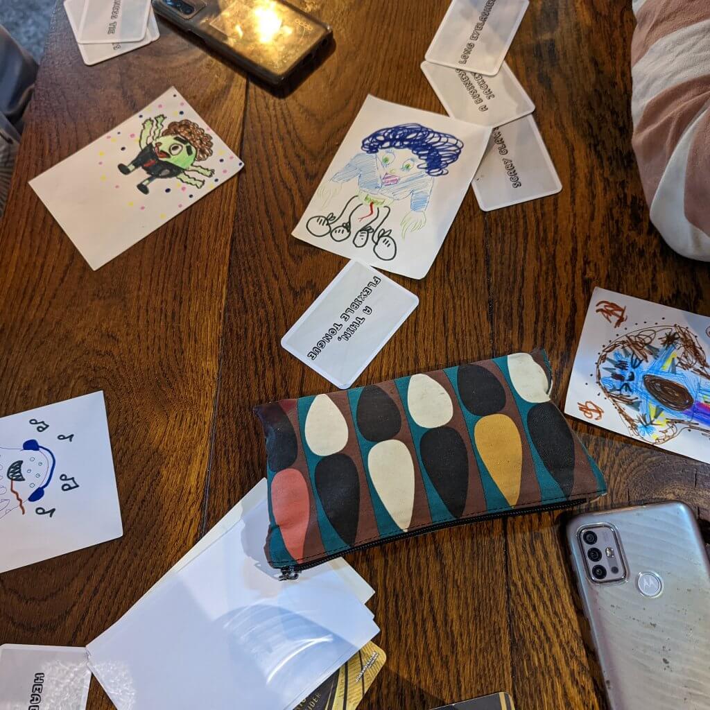 The drawing and compliments game. There are some cards with requirements on and some colourful creatures that have been drawn alongwith a pencilcase and a facedown phone .