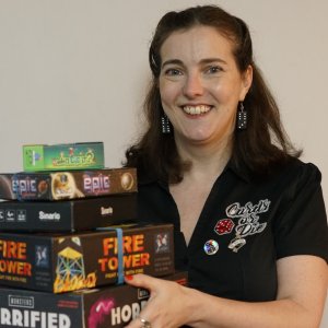 White woman with long dark hair. She is smiling. She holds a pile of boardgames in front of her.