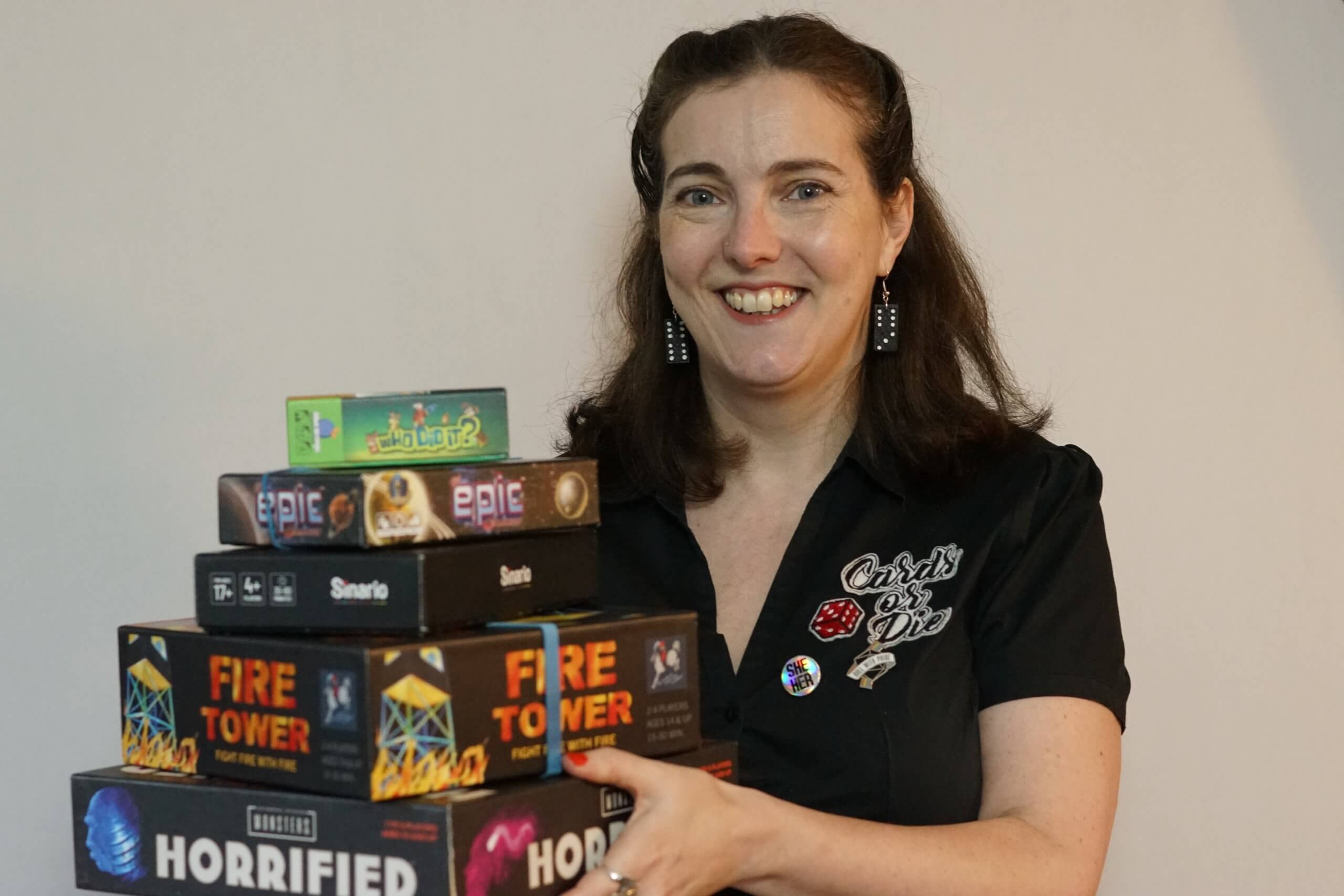 White woman with long dark hair. She is smiling. She holds a pile of boardgames in front of her.